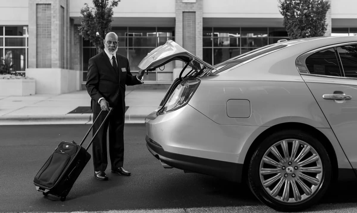 Professional chauffeur in a dark suit loading a roller suitcase into the trunk of a luxury sedan at airport curbside — flat-rate airport car service to Chicago O'Hare and Midway