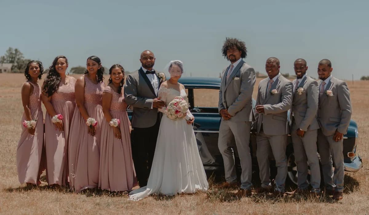 A full wedding party of bridesmaids and groomsmen flanking the couple beside a vintage car — coordinating vehicles for the wedding party