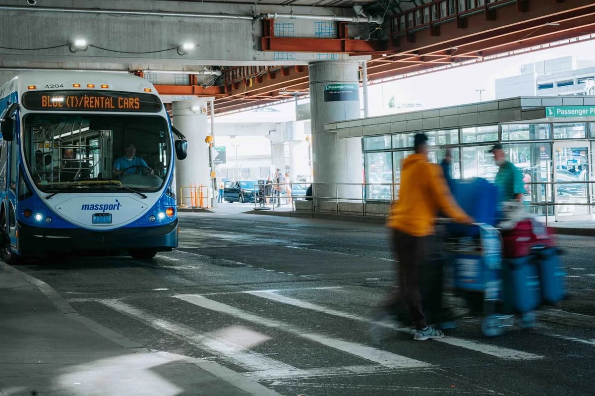 Airport curbside pickup zone with passenger-pickup signage, travelers crossing with luggage carts, and a shuttle under the terminal canopy