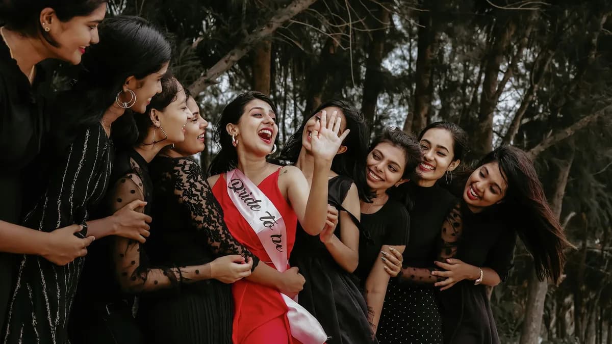 A bachelorette group of women surrounding a laughing bride-to-be in a red dress and sash, celebrating outdoors