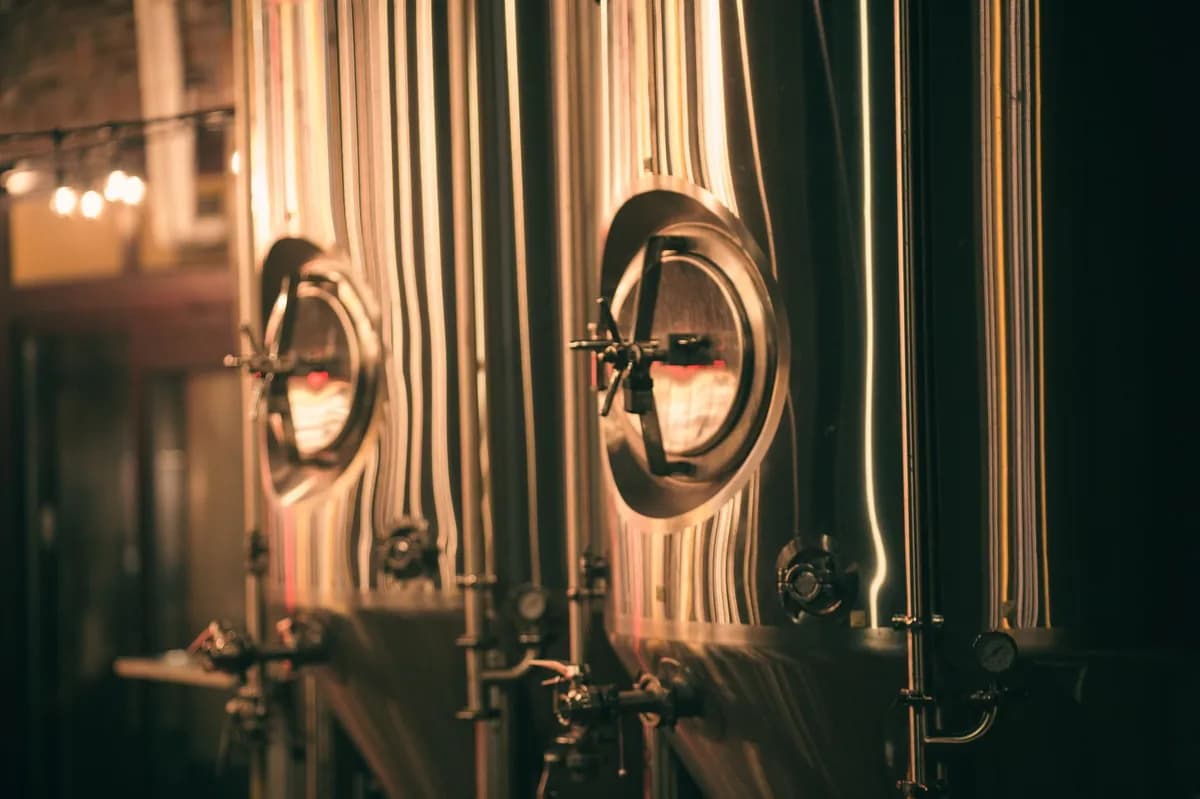 Gleaming stainless-steel fermentation tanks inside an industrial Pilsen-style craft brewery with warm string lights