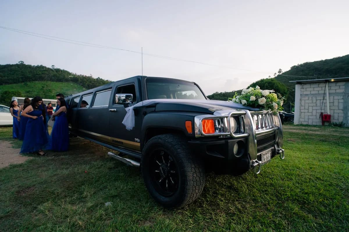 A formally dressed group in evening gowns beside a black Hummer stretch limousine at dusk — prom transportation